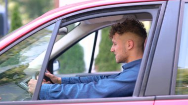 Close up of young handsome happy man sitting in red car listening to music and smiling to camera. Positive Caucasian male in auto in good mood looking at camera. Driver concept. Traveling in car