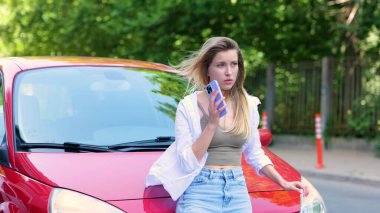 Portrait of beautiful young woman calling on smartphone while standing at her red car having problems with vehicle. Pretty upset worried woman calls tow truck on mobile phone. Breakdown on a road.