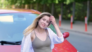 Close up portrait of young beautiful woman smiling and looking at camera in front of red car. Slow motion. Pretty Caucasian joyful female in good mood standing outdoors in street at her auto