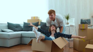 Happy young couple having fun in new bought apartment. Funny husband and wife playing on moving day. Excited adult man pushing cardboard box with his girlfriend sitting inside. Relocate in new home