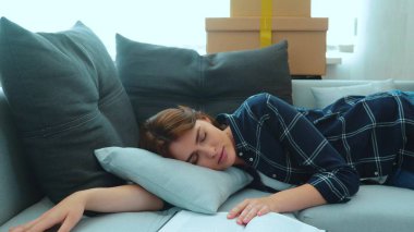 Beautiful young Caucasian woman lying on sofa in living room fallen asleep with book in new apartment with many cardboard boxes. Caring loving husband covers his wife with a blanket. Love concept