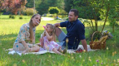 Happy young Caucasian man and woman couple spending weekend with child in park. Cheerful little girl playing with soap bubbles outside. Joyful family having picnic outdoor sitting on blanket in nature