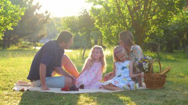 Happy family. Young cheerful parents spending time on nature with children sitting on a blanket. Handsome dad talking to cute daughter. Mom hugging child. Happy childhood. Parenthood. Weekend picnic