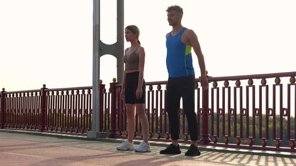 stock image Camera approaching young couple of man and woman in sportswear working out outside on the bridge. Caucasian sporty male and female doing exercise outdoor. Athletes doing squats. Sport activity.