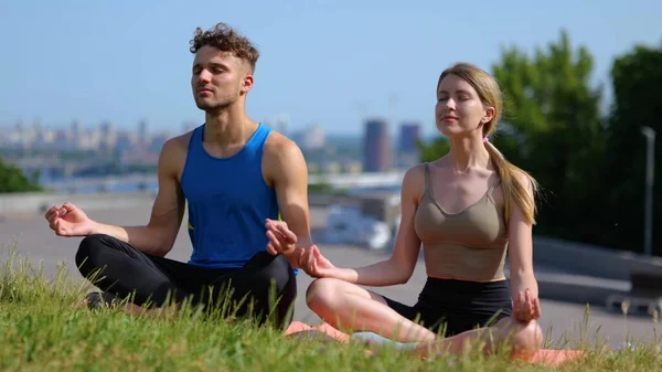 Young happy calm man and woman sitting in lotus pose meditating outside in city and breathing deeply. Male and female couple practicing yoga sitting on grass outdoors. Relaxation and harmony