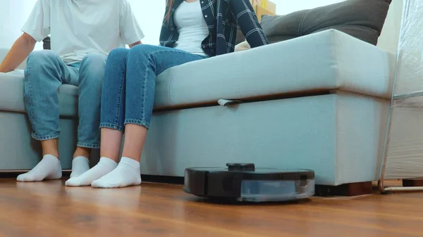 Close up of vacuum robot auto cleaning at home. Robot vacuuming in the modern living room. Couple raising their legs while sitting on sofa to let the vacuum cleaner pass on the floor. Home concept