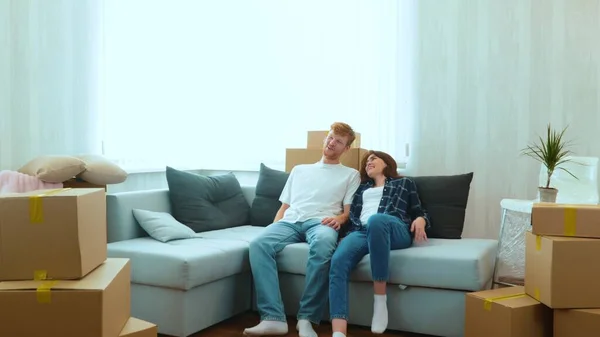 Cheerful Caucasian couple boyfriend and girlfriend moving in together in new house in good mood. Tired happy young married couple sitting down on sofa in new home with many boxes on moving day