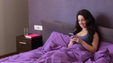 Mutlu bayan evde yatakta yatarken akıllı telefon kullanıyor. Cep telefonu tutan, ekrana bakan ve daktilo yazan genç bir kadın. Teknoloji, internet, iletişim konsepti. Yavaş çekim