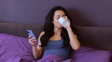 Gülümseyen genç bayan sabah yatak odasında kahvesini yudumlarken akıllı telefon kullanıyor ve internette sohbet ediyor. Yaşam tarzı konsepti. Yavaş çekim