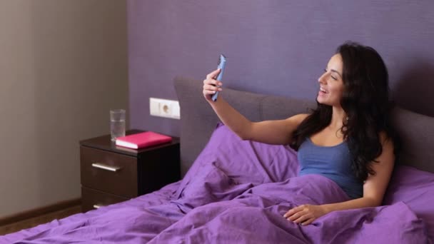 스마트폰을 사용하며 침대에 행복하고 아름다운 아가씨 즐거운 비디오 전화를 집에서 — 비디오