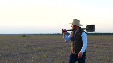 Hasır şapkalı mutlu erkek işçi tarlada yürüyor ve hasatını inceliyor. Kürek tutan çiftçi. Tarım sektörü konsepti. Gerçek zamanlı.