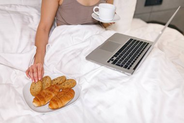 Genç bayan sabah bir fincan sıcak içecekle yatakta oturuyor. Kadın kurabiye yiyecek, dizüstü bilgisayarı bacağında.