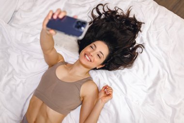 Yatakta yatan ve selfie çekerken cep telefonu tutan gülümseyen bayanın en iyi görüntüsü. Eğlence konsepti