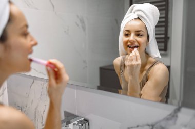 Gülümseyen güzel bayan banyoda aynanın önünde dikilirken dudaklarına çıplak ruj sürüyor. Makyaj, sabah ve yaşam tarzı rutini