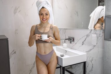 Gülümseyen güzel bayan banyoda dikilirken sıcak içkinin tadını çıkarıyor. Beyaz havluyla kahve içen bir kadın.