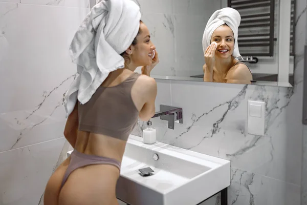 鏡を見ながらシャワー後の朝のスキンケアルーチンを行う幸せな若い女性は バスルームに立っている 美容とトリートメントのコンセプト — ストック写真
