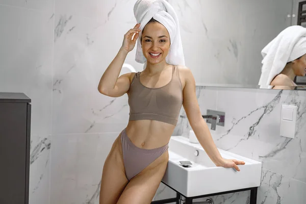 一边在浴缸里摆姿势一边微笑着穿着内衣的年轻女人 一边摸着毛巾放在头上 快乐的女人站在浴室里 — 图库照片