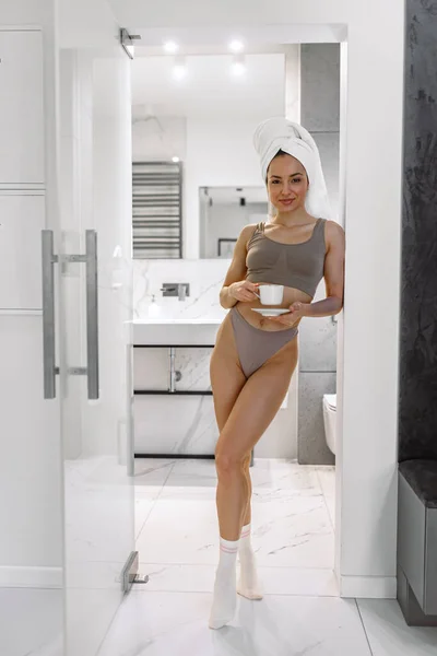 穿着内衣微笑的女人站在浴室里 举着一杯咖啡 摆出一副相机的样子 早上在家的例行公事 — 图库照片