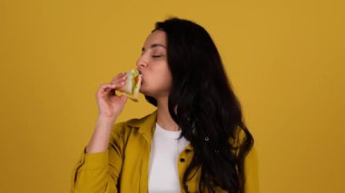 Sandviç yerken stüdyoda poz veren genç bir kadın. Sarı arka planda izole edilmiş. Kopya alanı, insanlar, yaşam tarzı, abur cubur konsepti. Gerçek zamanlı.