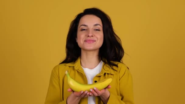 黄色の背景に隔離されたスタジオでポーズをとっている間 1つのバナナを持っている幸せなかなり女性 健康的で新鮮な食べ物の概念 リアルタイム — ストック動画
