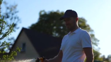 Gülümseyen bahçıvan, sulama bitkileri için fıskiye hortumunu tutuyor. Köknar ağacını önemseyen adam. Bahçe işleri konsepti. Gerçek zamanlı.