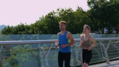 Mutlu çift köprüde koşarken antrenmanın tadını çıkarıyor. Bir şişe su tutan adam. Egzersiz, koşu ve spor konsepti. Gerçek zamanlı.