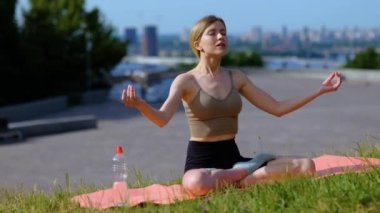 Spor kıyafetli güzel bir kadın şehirde meditasyon yaparken yoga minderinde oturuyor. Sakinlik ve meditasyon kavramı. Gerçek zamanlı.