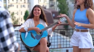 Arkadaşları çatıdaki partide dans ederken gülümseyen çekici bayan gitar çalıyor. Yaşam tarzı, arkadaşlık, eğlence, açık parti konsepti. Gerçek zamanlı.