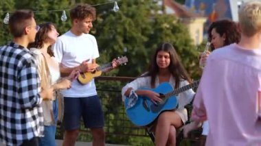 Neşeli bir kadın gitar çalarken bir adam çatıdaki partide ukulele kullanıyor, arkadaşları dans ediyor, bira tutuyor. Yaşam tarzı, arkadaşlık, eğlence, açık parti konsepti. Gerçek zamanlı.