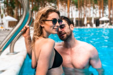 Açık havuzda genç bir adamla birlikte duran bikinili sevimli sarışın kadın. Romantizm ve yaz tatili kavramı