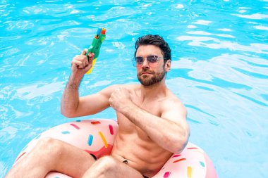 Yüzme havuzunda oyuncak su tabancası tutarken şişme halkada oturan güneş gözlüklü sakallı adam. Yaz zamanı, tatiller, yaşam tarzı konsepti