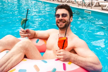 Güneş gözlüklü, gülümseyen, hafta sonunun tadını çıkaran ve havuzda iki kokteyl içen adam. Yaz zamanı, tatiller, yaşam tarzı konsepti.