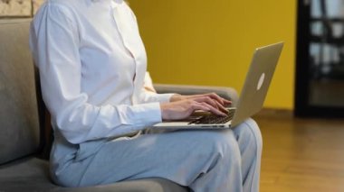 Sarı duvarlı çağdaş ofis, gerçek zamanlı video. Beyaz takım elbiseli, beyaz tişörtlü, siyah, uzun saçlı beyaz bir kadın kanepeye oturmuş laptopunda yazı yazıyor ve kameraya bakıyor..