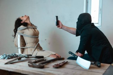 Siyah elbiseli bir adam akıllı telefon tutuyor ve bodrumda iple bağlanmış kaçırılmış bir kadının videosunu çekiyor. Suç, adam kaçırma, şiddet kavramı