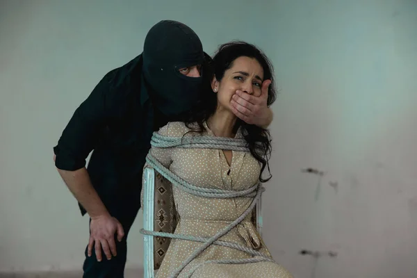 Kidnapper in a black balaclava covering mouth of frightened lady by his hand. Female tied on a chair with a rope. Crime, kidnapping, violence concept