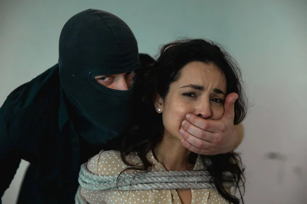 Bandit in a black balaclava covering the woman mouth with his hand, lady sitting on a chair tied with a rope. Crime, kidnapping, violence concept