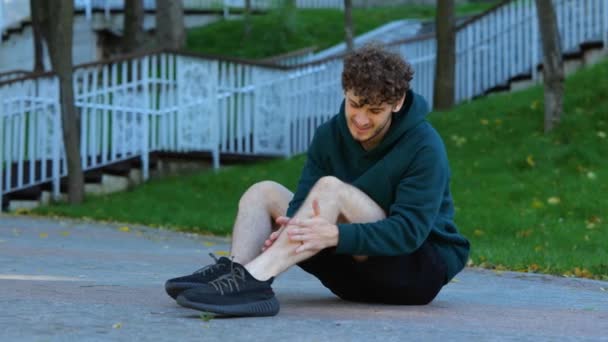 若い白人男性が地面に座り 緑の公園の筋肉を伸ばしました スポーツマンは彼の足に強い痛みを感じ 働く前に膝と筋肉をマッサージする — ストック動画