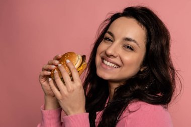 Çekici bir sandviçle pembe arka planda kameraya poz veren hoş bir genç bayanın portresi. İştah ve favori fast food konsepti