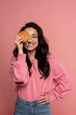 Stüdyo fotoğraf çekimi sırasında pembe arka planda poz verirken bir gözünü lezzetli bir çizburgerle kaplayan mutlu güzel bayan.