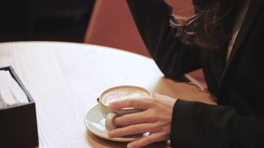 Young Caucasian girl with long dark hair drinking cappuccino from a white coffee cup inside a restaurant, girl is sitting at a round wooden table and looking at the screen of her smartphone.