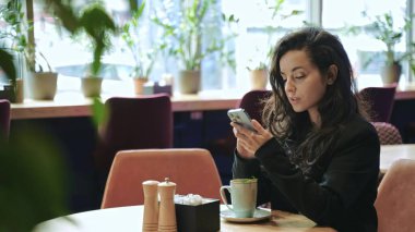 Yakın çekim, yavaş çekim. Kafkasyalı güzel iş kadını bir sürü yeşil bitkiyle modern restoranın içinde oturuyor, naneli sıcak çay içiyor ve telefonuna kayıyor ve daktilo çekiyor..
