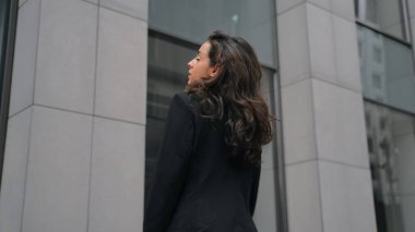 Back view, close up, slow motion. Woman with long dark hair and black suit turns to the camera, smiling, and putting her hands on her chest. Outdoors, grey office modern buildings, city center.