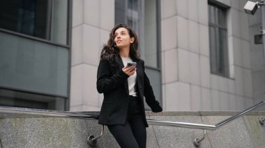 Outdoors, remote work, slow motion. Young Caucasian businesswoman wearing black jacket standing next to office buildings, smiling, looking around, and scrolling her smartphone. Outdoors, remote work.