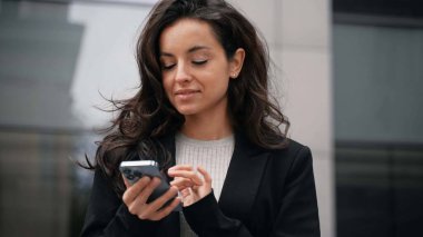 Young girl wearing office style jacket standing downtown. Caucasian businesswoman standing next to office buildings, smiling, looking around, touching her dark long hair, scrolling her smartphone.
