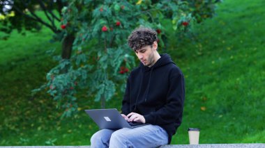 Adam dizüstü bilgisayarla çalışıyor ve parkta oturmuş kahve içerken daktilo yazıyor. Sonbahar sezonu. Eğitim, çalışma konsepti. Yavaş çekim