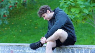 Adam yeşil parkta bacağına masaj yapıyor beton bankta oturuyor. Sporcu ayak ve ayak bileğinde güçlü bir ağrı hissediyor. Kahverengi kıvırcık saçlı, koşudan sonra bacağını kolalayan yakışıklı adam..