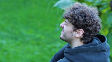 Kenar manzaralı genç beyaz adam yeşil parkta bacağına masaj yapıyor beton bankta oturuyor. Siyah noktalı şortlu sporcu, kapüşonlu, spor ayakkabılı rahatlayan, bacağına masaj yapan, bacağında ağrı hisseden..