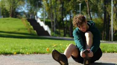 Genç beyaz bir adam güneşli bir yaz gününde yerde oturup parkta ayağına masaj yapıyor. Şiddetli acı çeken bir adamın yüzü. Açık hava aktiviteleri. Sporcunun bacağında şiddetli ağrı var..