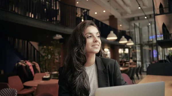 Close up, slow motion. Beautiful businesswoman working on her computer inside of cafe. Girl working remotely, she is enjoying and looks successful and happy. Woman smiling and waving her hand.
