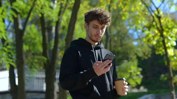 街の公園を歩きながらコーヒーを楽しんでいるハッピーな男が 携帯電話で話している ライフスタイル 秋歩きのコンセプト スローモーション — ストック写真